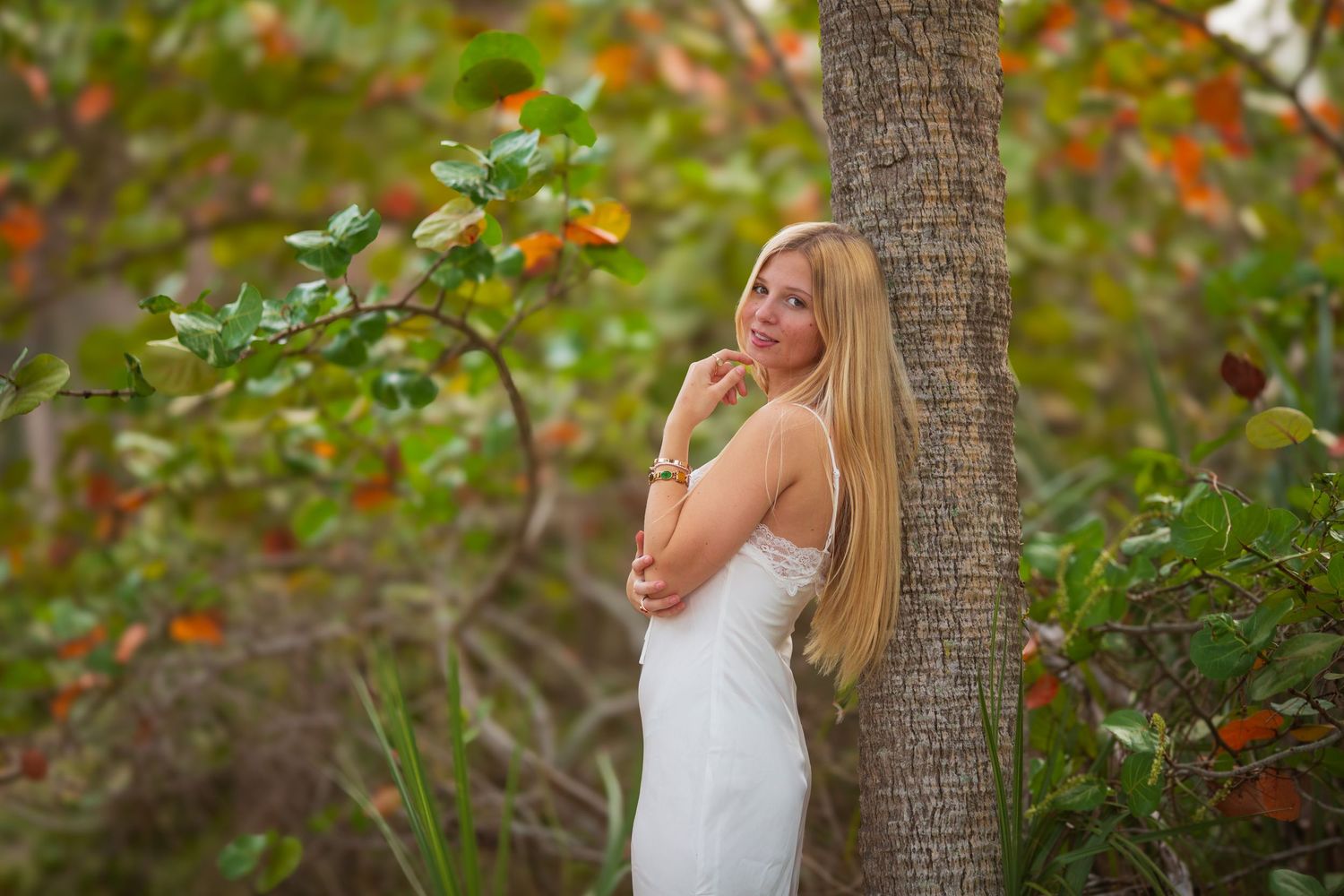 Blonde woman in white dress leaning against tropical tree surrounded by lush green and orange foliage outdoors.