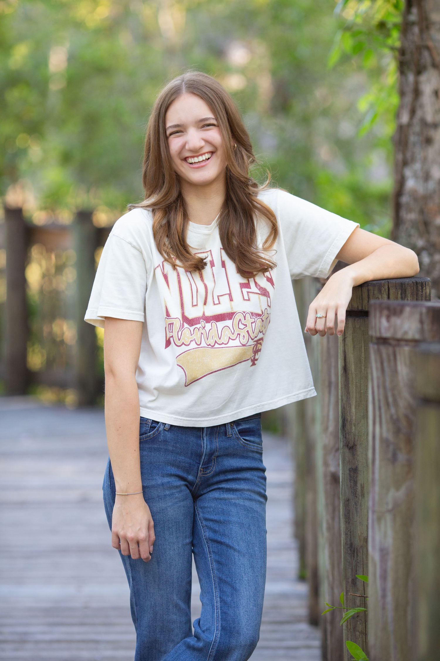 Smiling teenage girl in white graphic tee and jeans leaning on wooden fence on outdoor boardwalk.