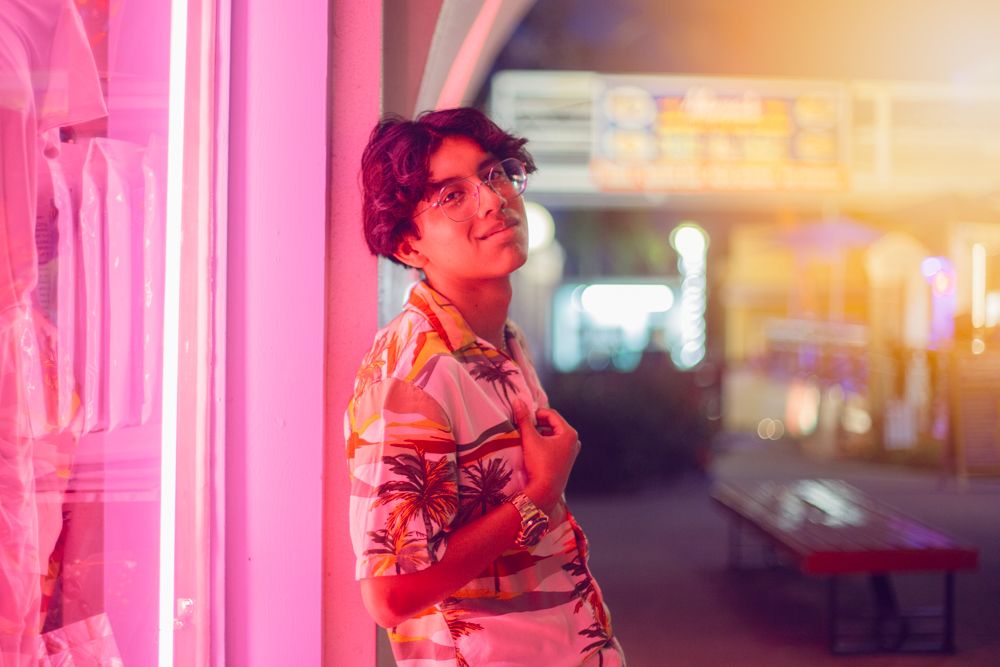 Young person in floral shirt posing by pink neon lights at night in urban setting.