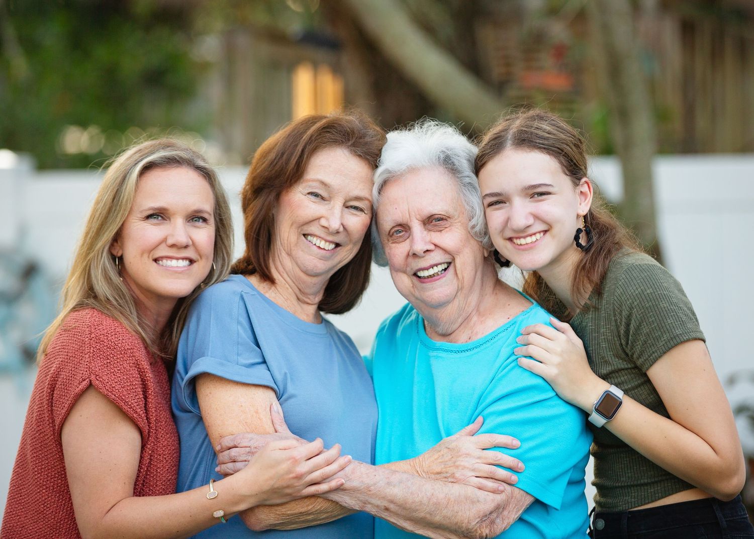Four women of different generations smile and embrace outdoors.