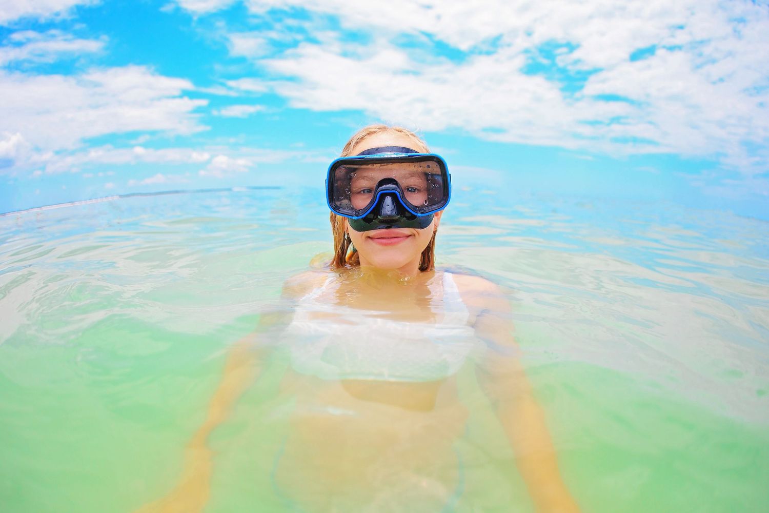 A snorkeler wearing goggles floats in crystal clear turquoise water under bright blue skies.