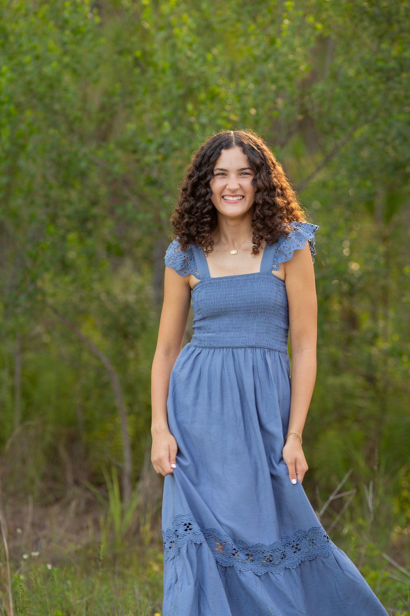 Young woman with curly hair smiling outdoors in a blue midi dress with lace trim, surrounded by lush green foliage.