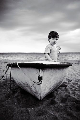 Artistic black and white photo of child sitting in old boat on sandy beach.