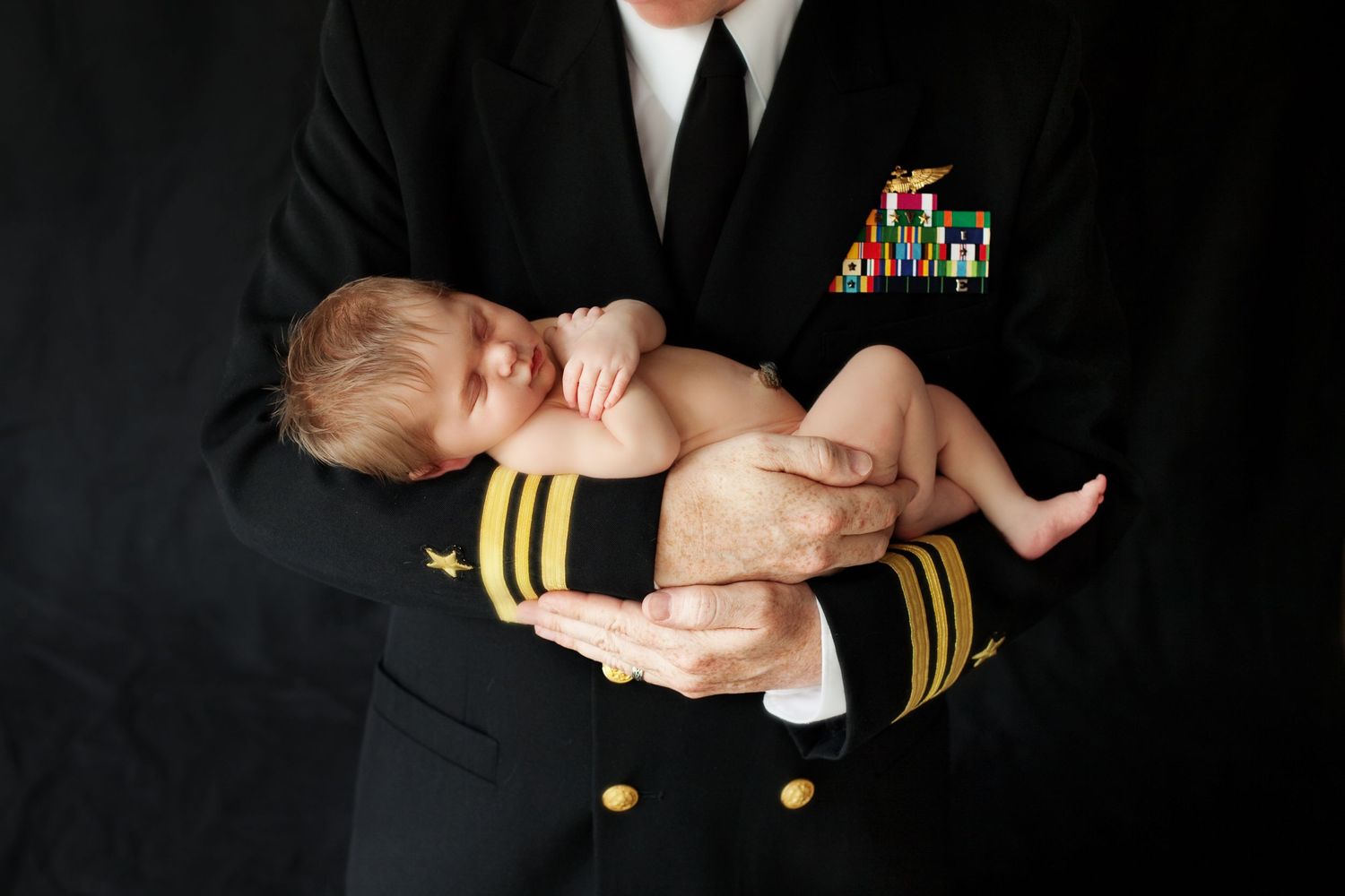 Newborn baby sleeping peacefully in military officer's uniformed arms against dark background.