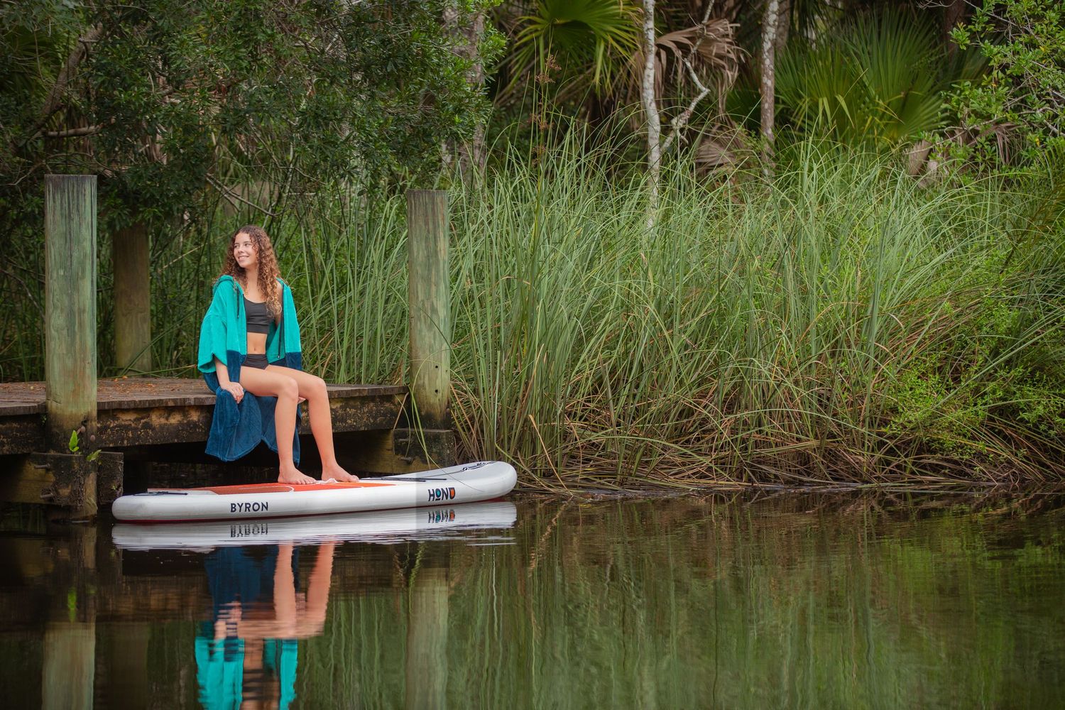 Woman in teal wrap sits on dock beside inflatable paddleboard, surrounded by lush green reeds and calm water.