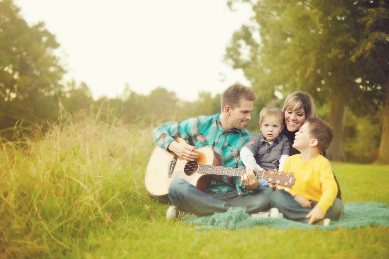 Family sitting on blanket in sunny field while playing acoustic guitar together.