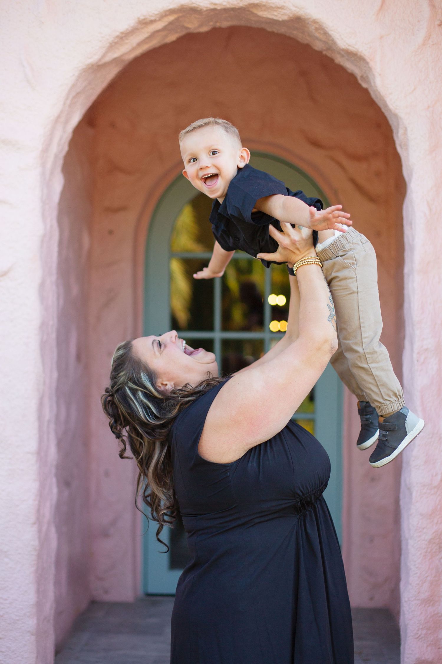 A young child is lifted playfully into the air by an adult in front of an arched entryway with a turquoise door.