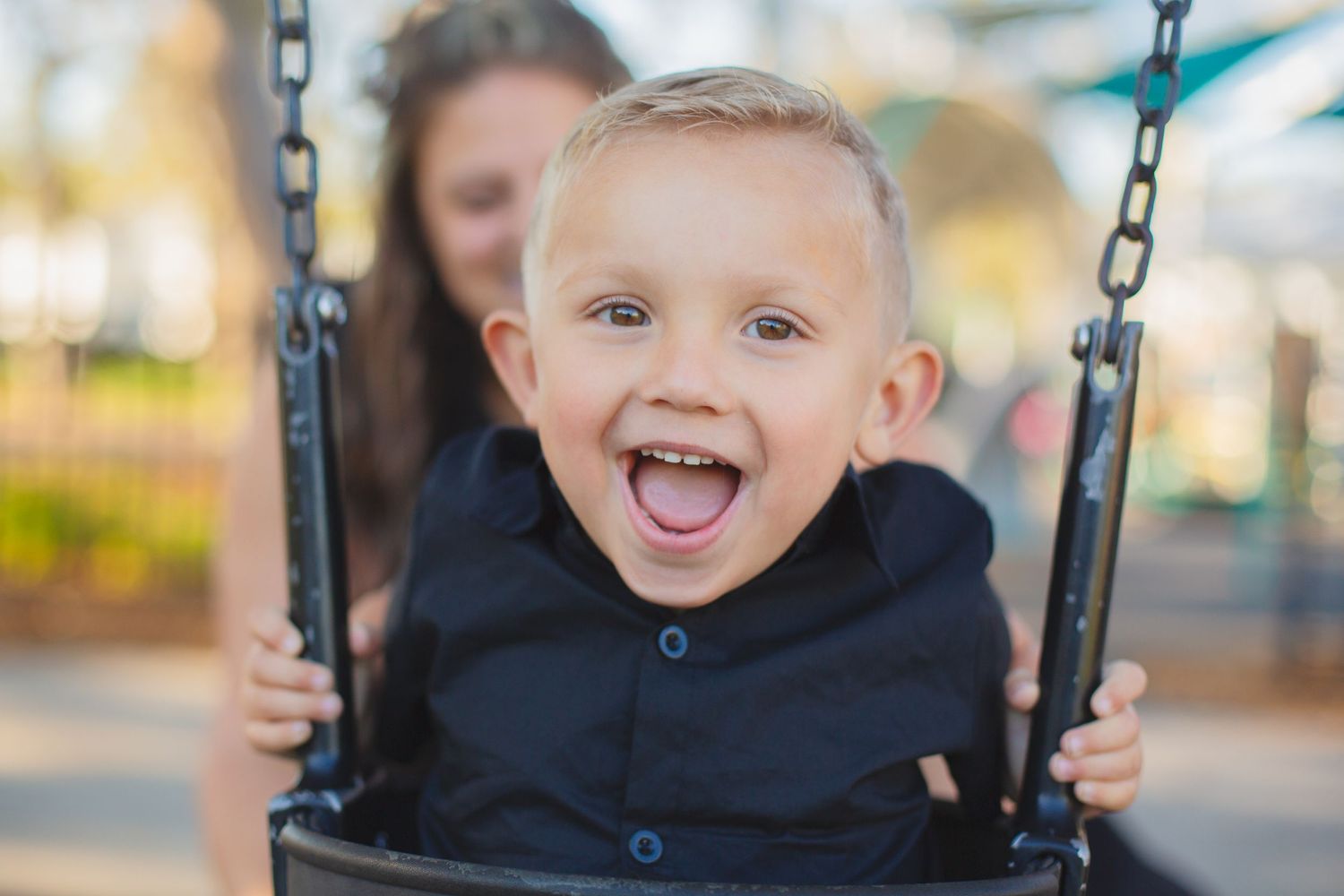 Happy child in black hoodie laughing with pure joy while swinging at a playground on a sunny day.