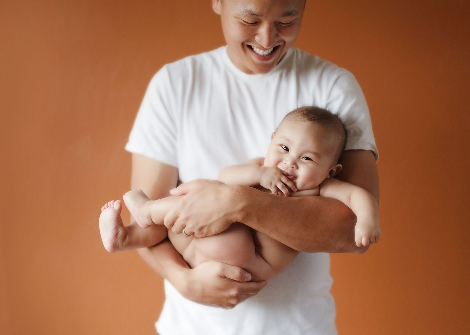 Parent in white shirt holding laughing baby against orange background.