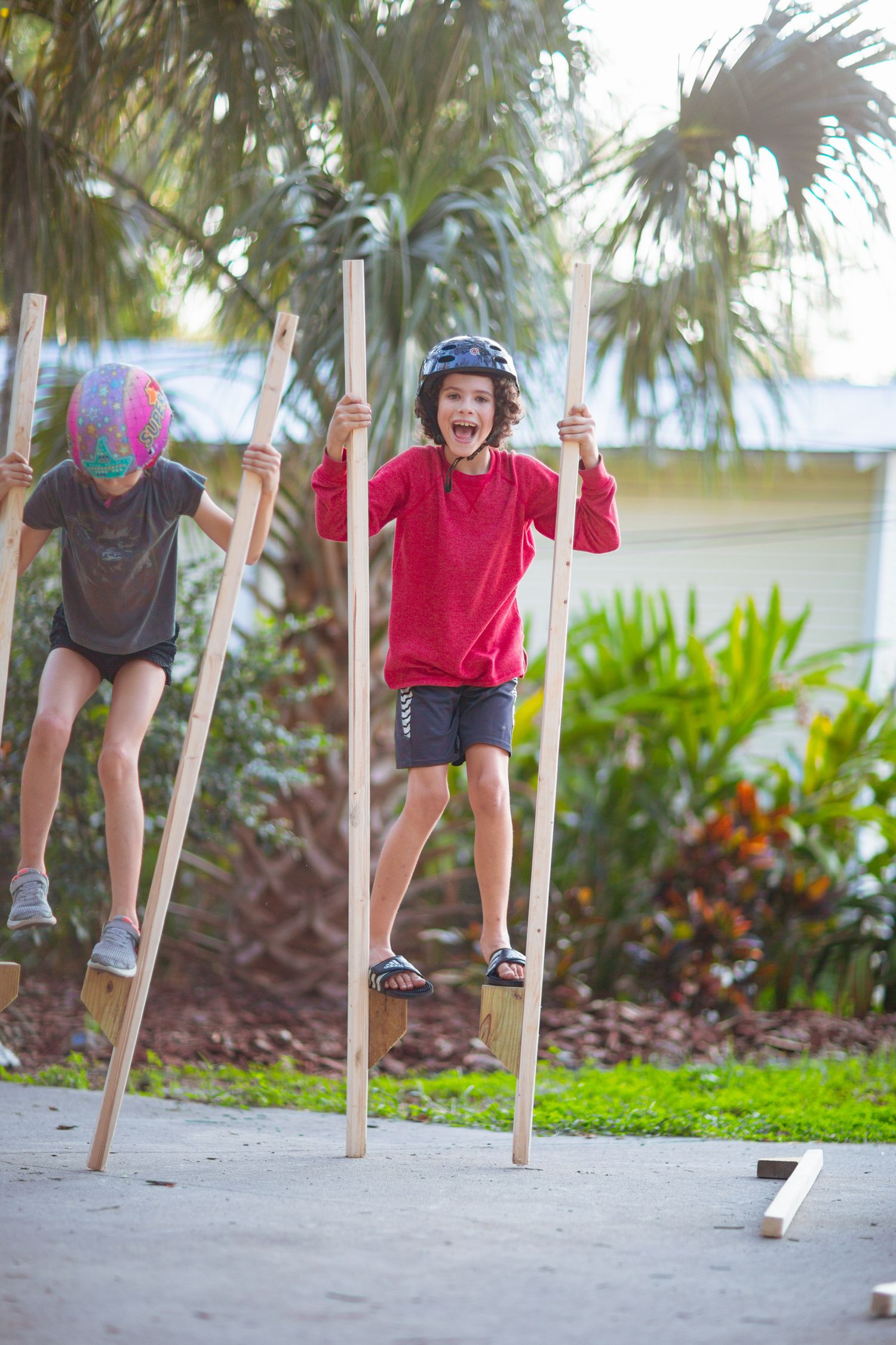Children walking on wooden stilts outdoors near palm trees.