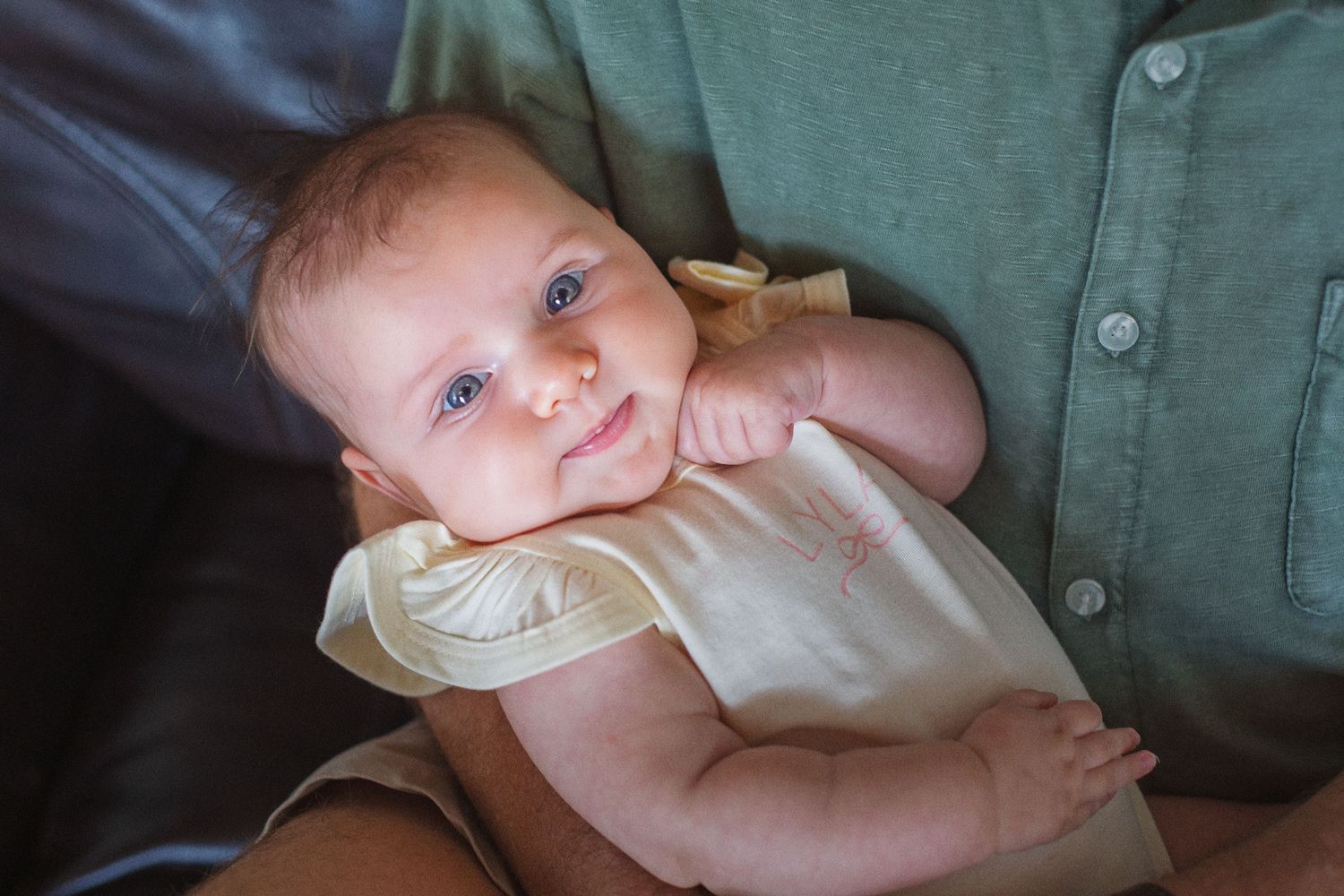 A smiling baby with blue eyes wearing a yellow onesie being held by an adult in a green shirt.