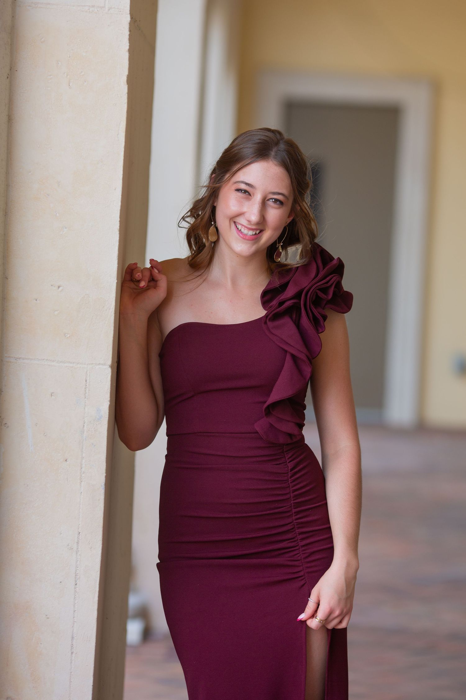 Young woman smiling in elegant burgundy one-shoulder ruffle gown, posing gracefully against a stone column.