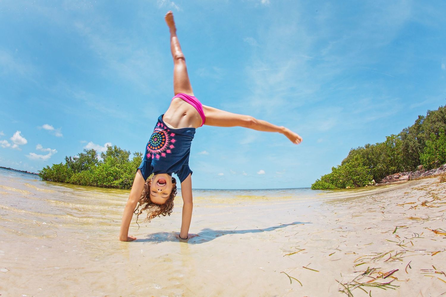 A young gymnast performs a cartwheel on a sandy beach against blue sky and ocean.