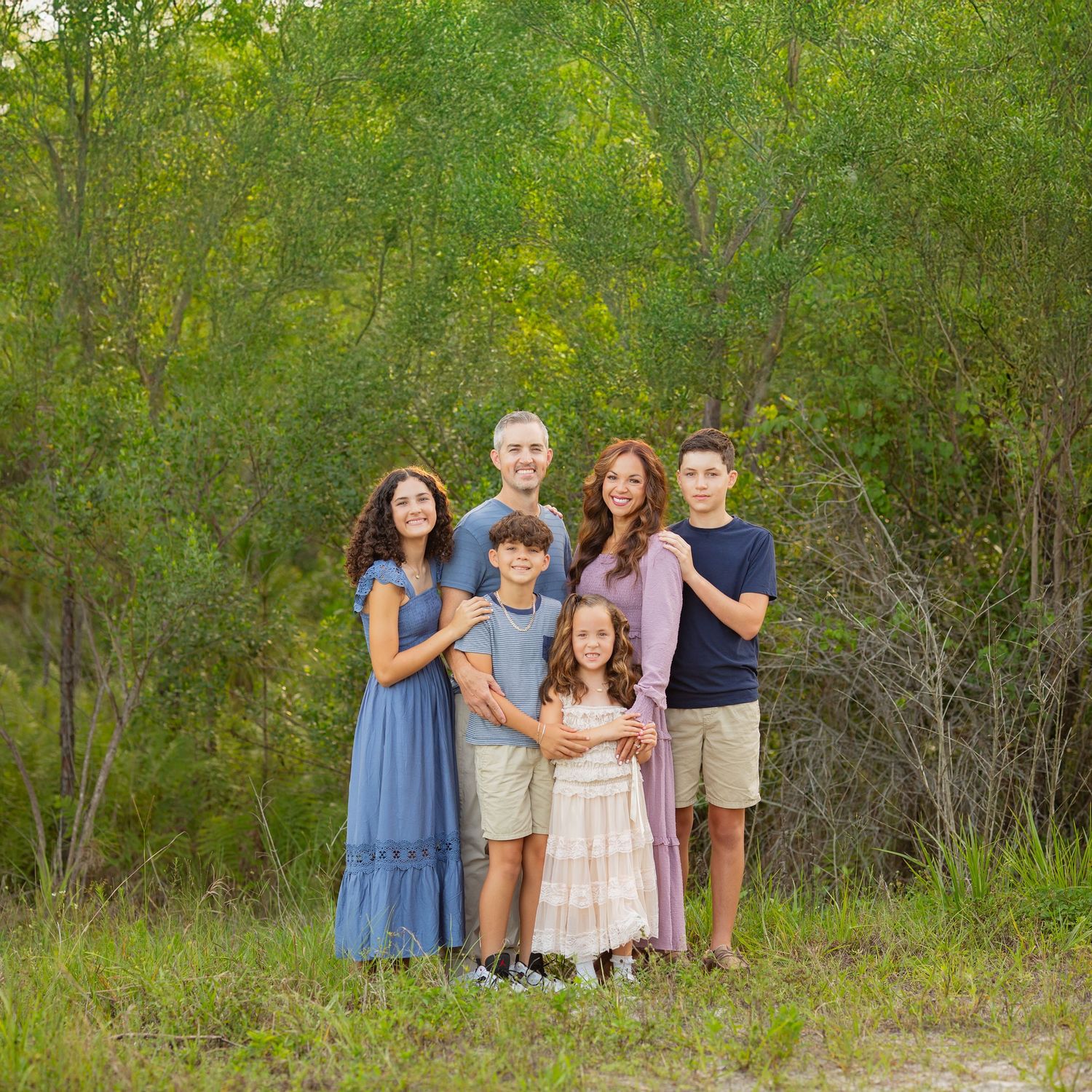 Family of six posing outdoors in coordinated blue and neutral outfits surrounded by lush green trees.