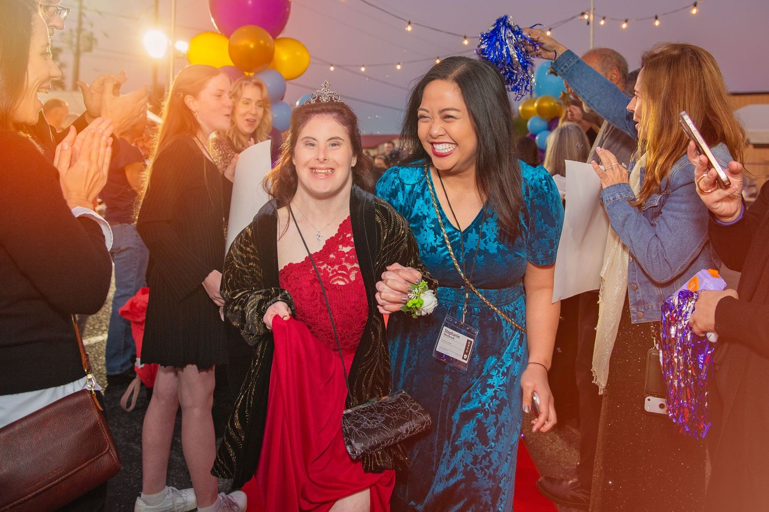 Two women in formal dresses smile and walk the red carpet at a festive evening event with balloons and guests.