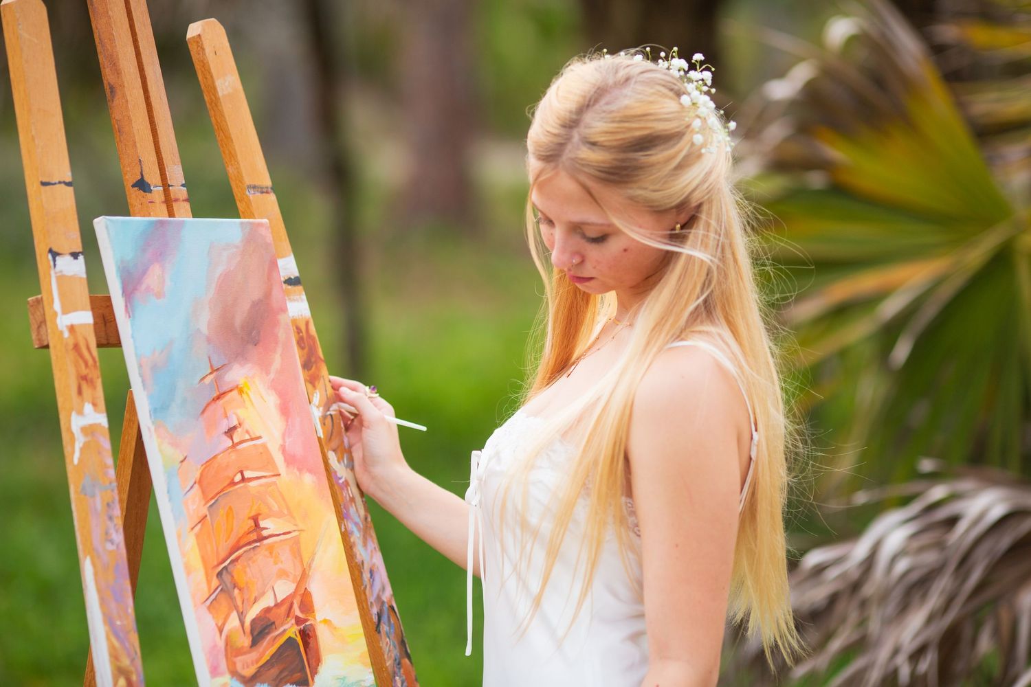 Blonde woman in white dress painting on easel outdoors, surrounded by lush green trees in a serene garden setting.