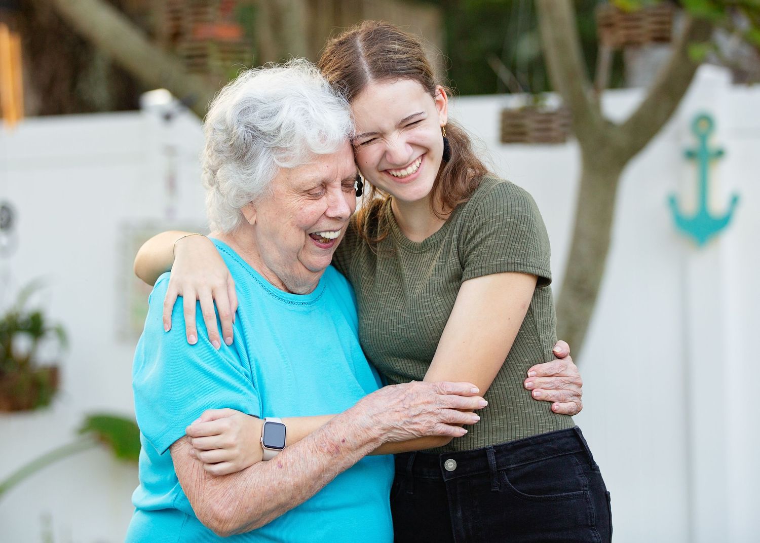 Two women share a close embrace and laugh together beside a white fence with an anchor decoration.
