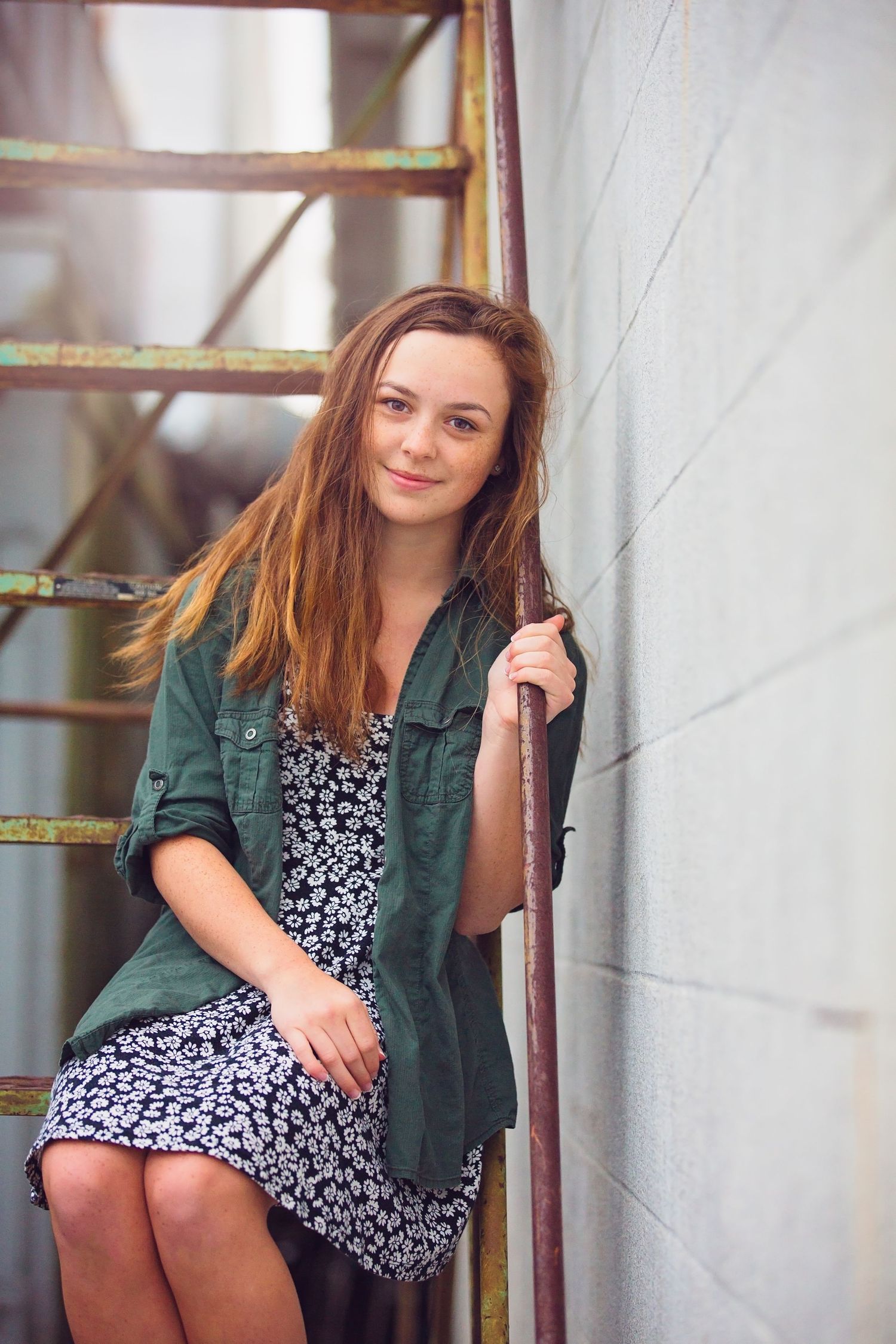 Portrait of someone in a green jacket and floral dress sitting on metal stairs.