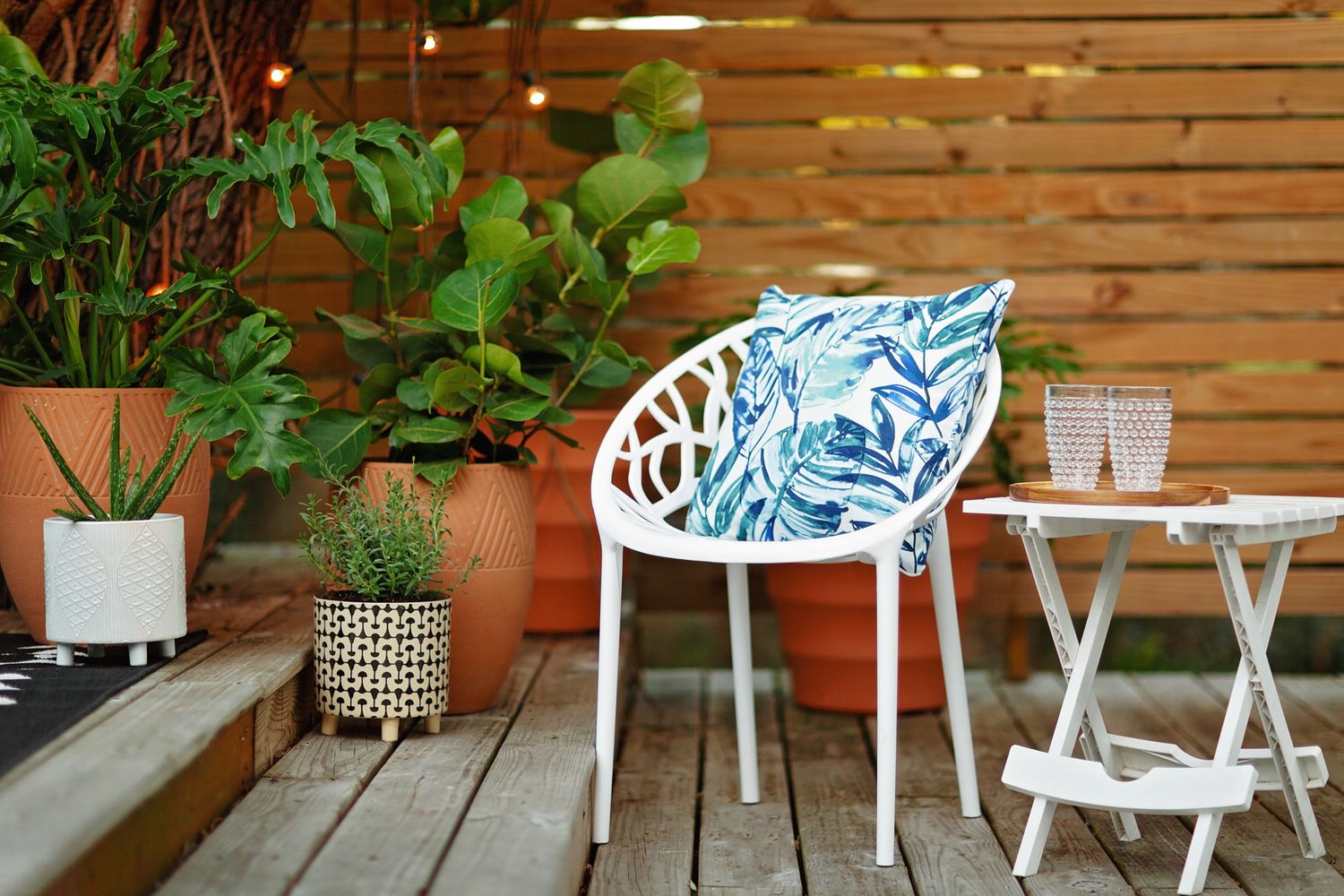 Modern white outdoor patio furniture with blue cushions arranged against wooden fence with potted plants.