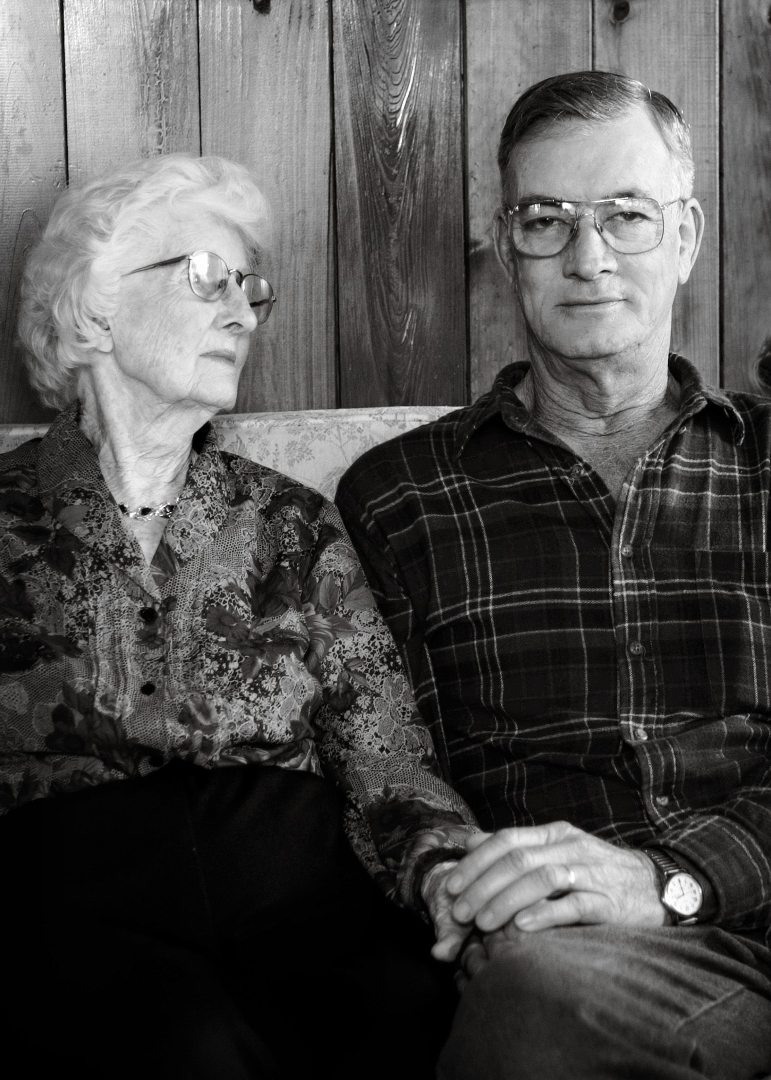 Black and white portrait of elderly couple sitting together against wooden wall.