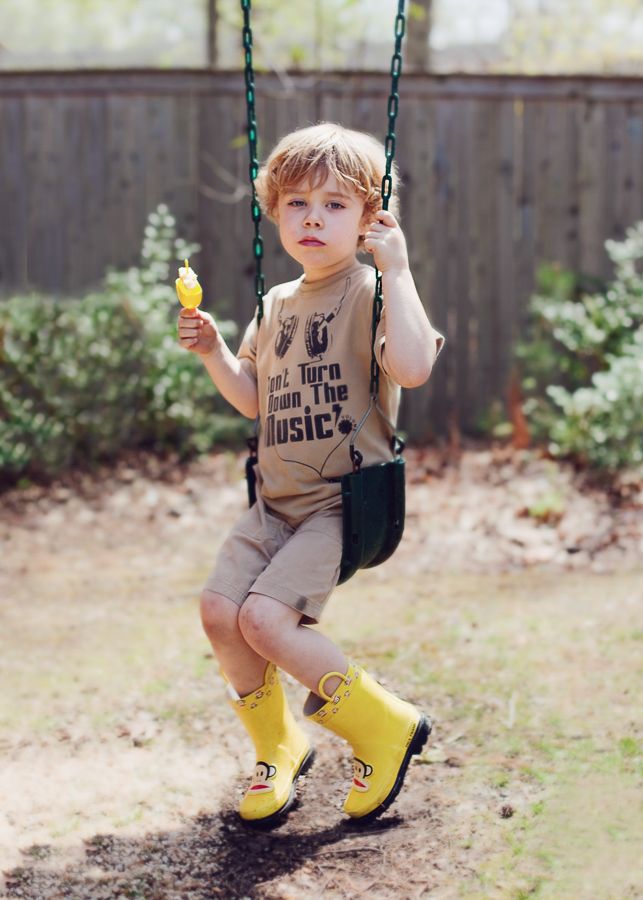 Child on swing wearing yellow boots and holding a snack in backyard garden.