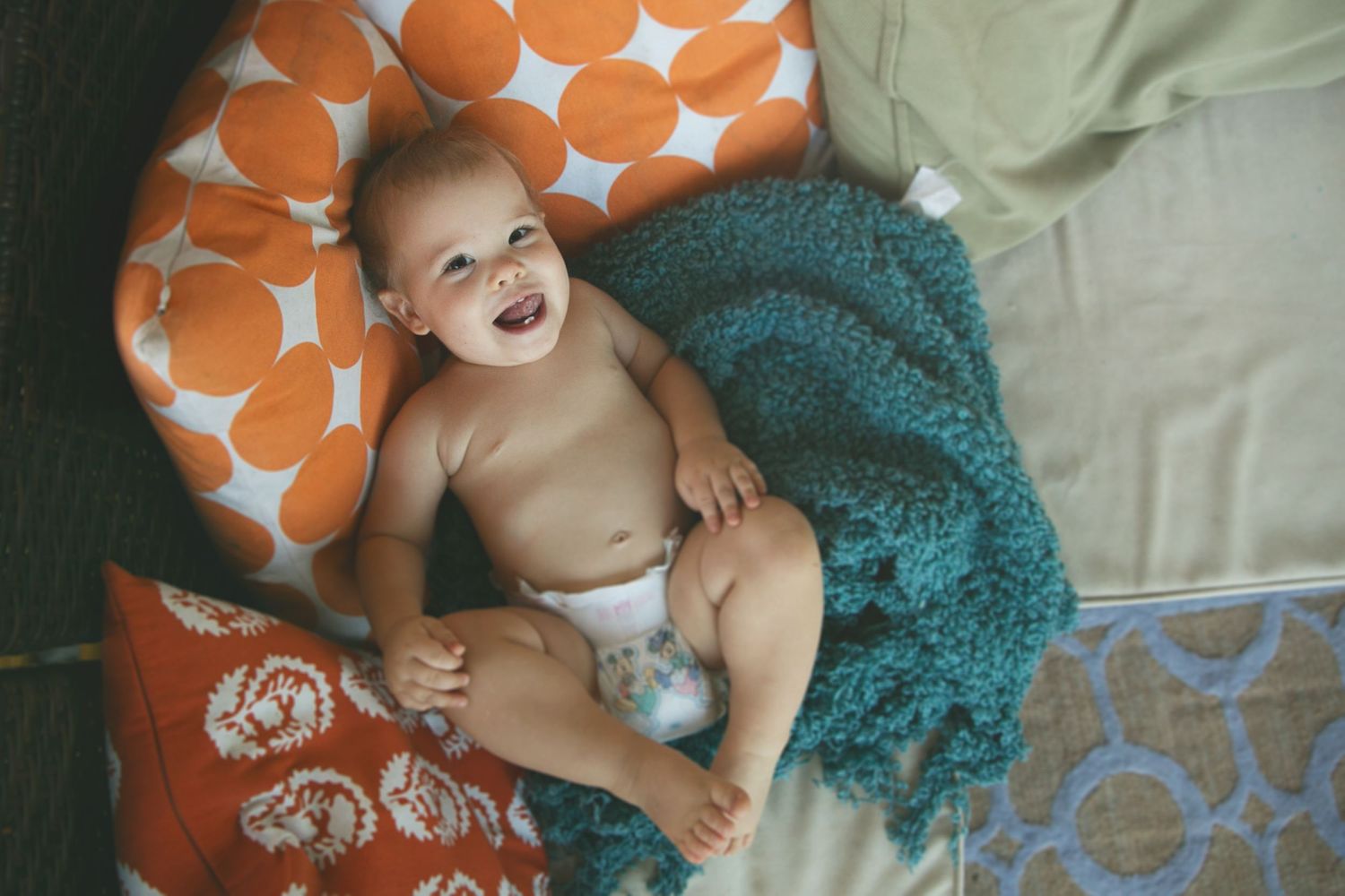 A happy baby lying on colorful pillows with orange polka dots and a teal blanket.