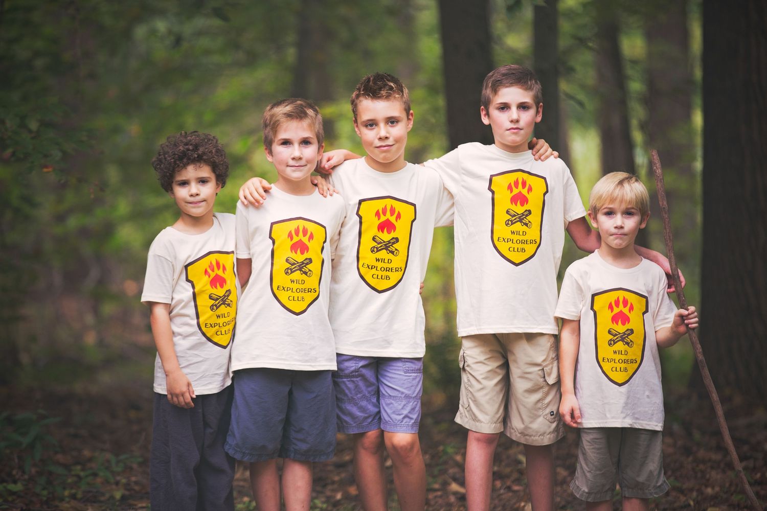 Children wearing matching shield logo t-shirts standing together in a forest.
