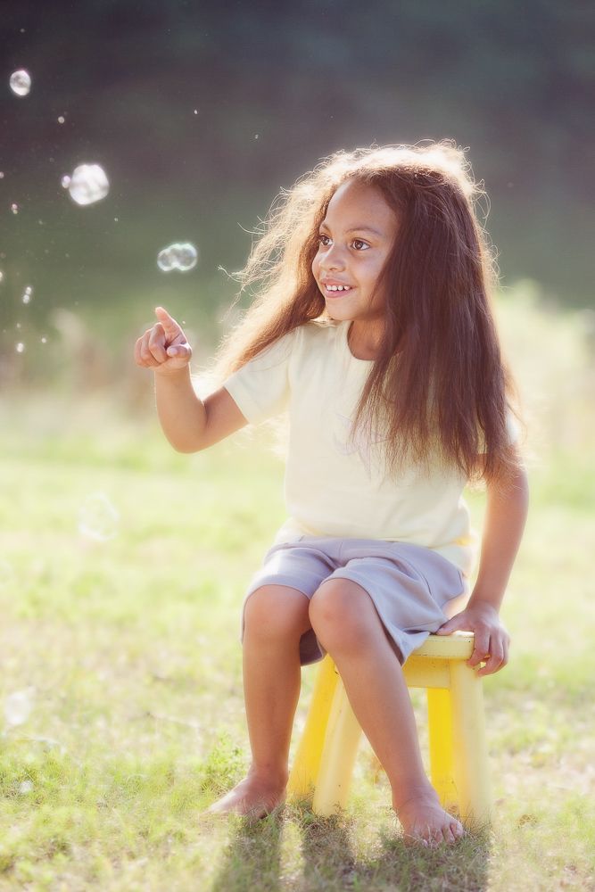 Young child with long hair sitting on yellow stool outdoors in magical sunlit setting with bubbles.