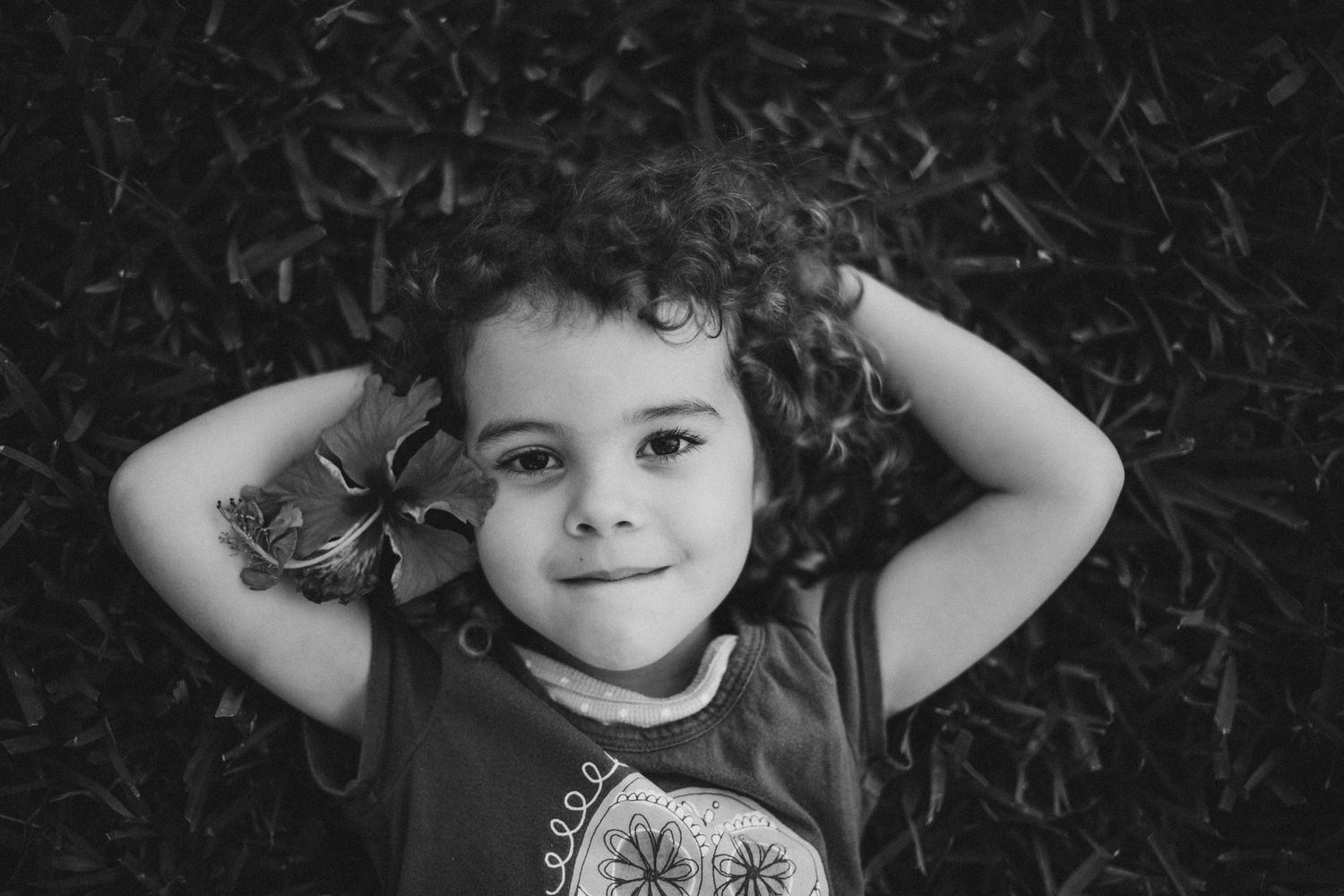 Black and white portrait of child with curly hair lying on ground looking up at camera with playful expression.