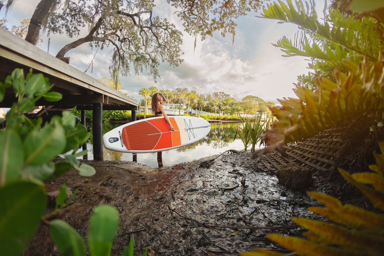 Woman carrying a red and white paddleboard near a tropical waterway surrounded by lush greenery.