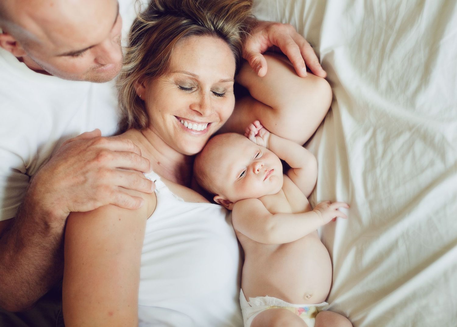 Family enjoying tender moment with newborn baby lying together on white bedding.