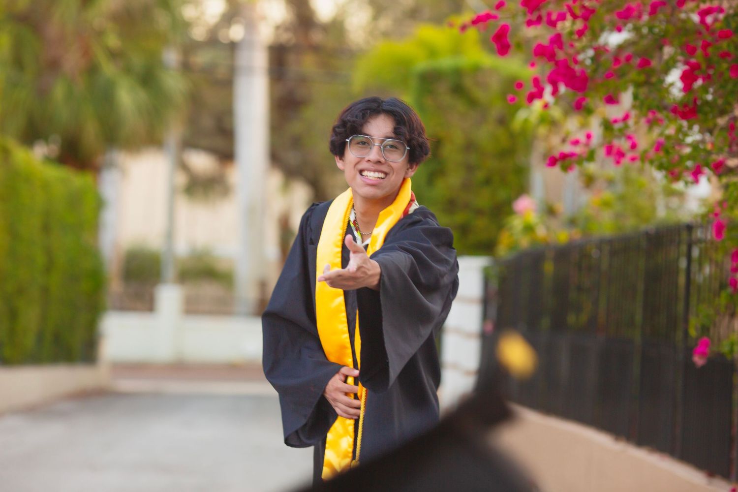 Graduate in black gown and gold sash smiling outdoors near pink flowers and green trees.