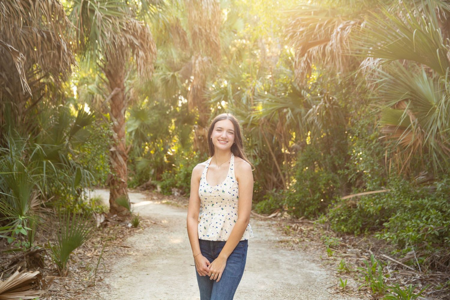 Young woman smiling on a sandy path surrounded by lush tropical palm trees in bright sunlight.