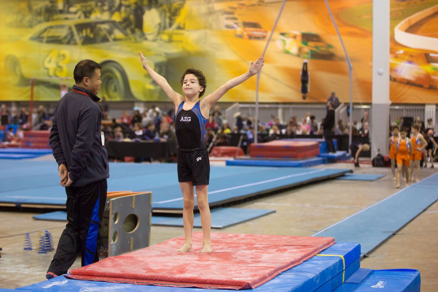 Young gymnast stands on red mat with coach in indoor gymnastics facility.