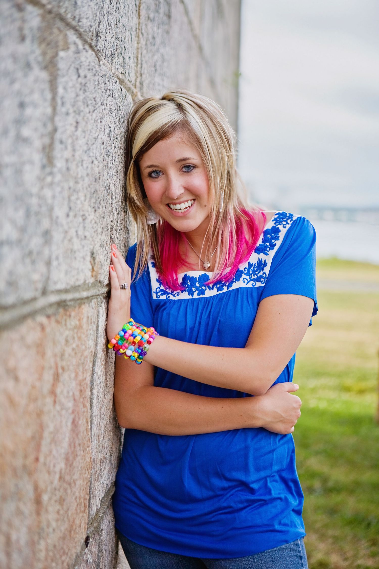 High school senior girl wearing a bright blue embroidered top with colorful bracelet poses against stone wall outdoors.