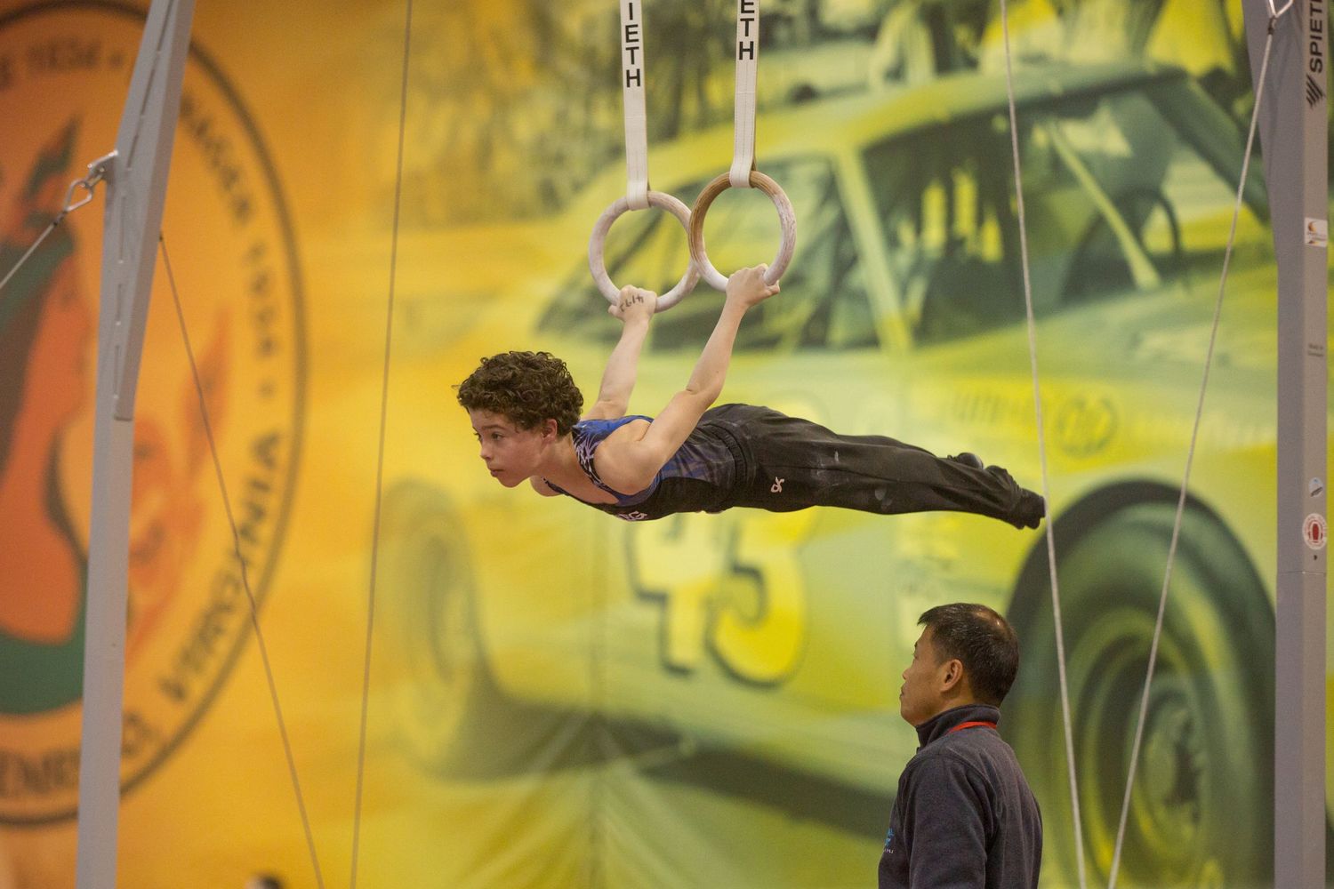 Person performing aerial gymnastics against a yellow backdrop with a spotter below.