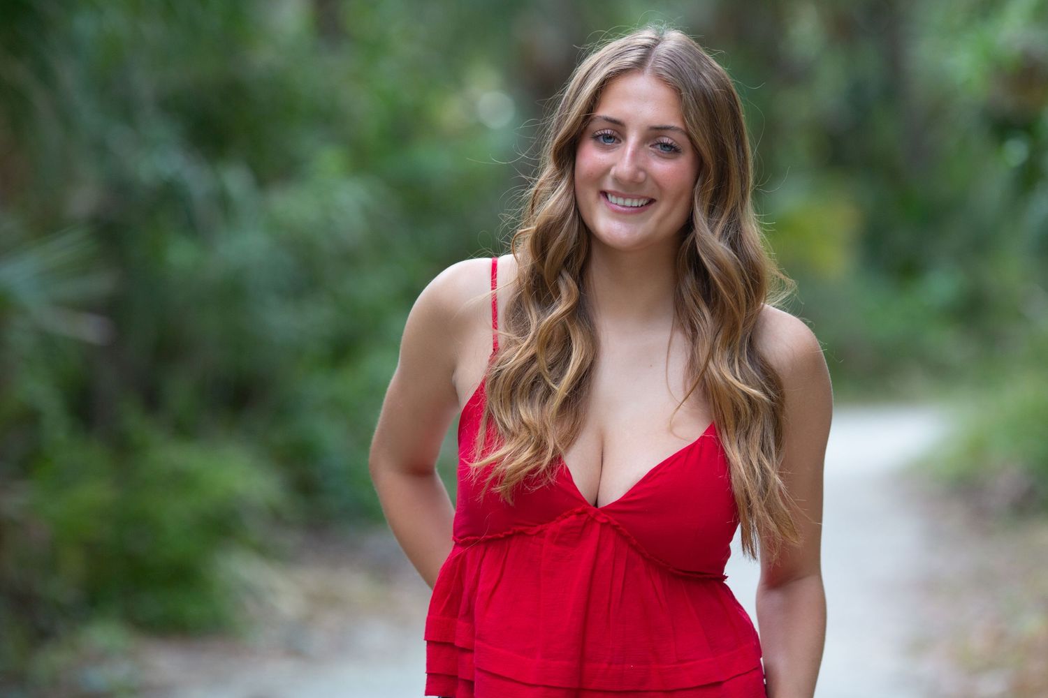 A person in a red top smiles while standing on a nature path surrounded by greenery.