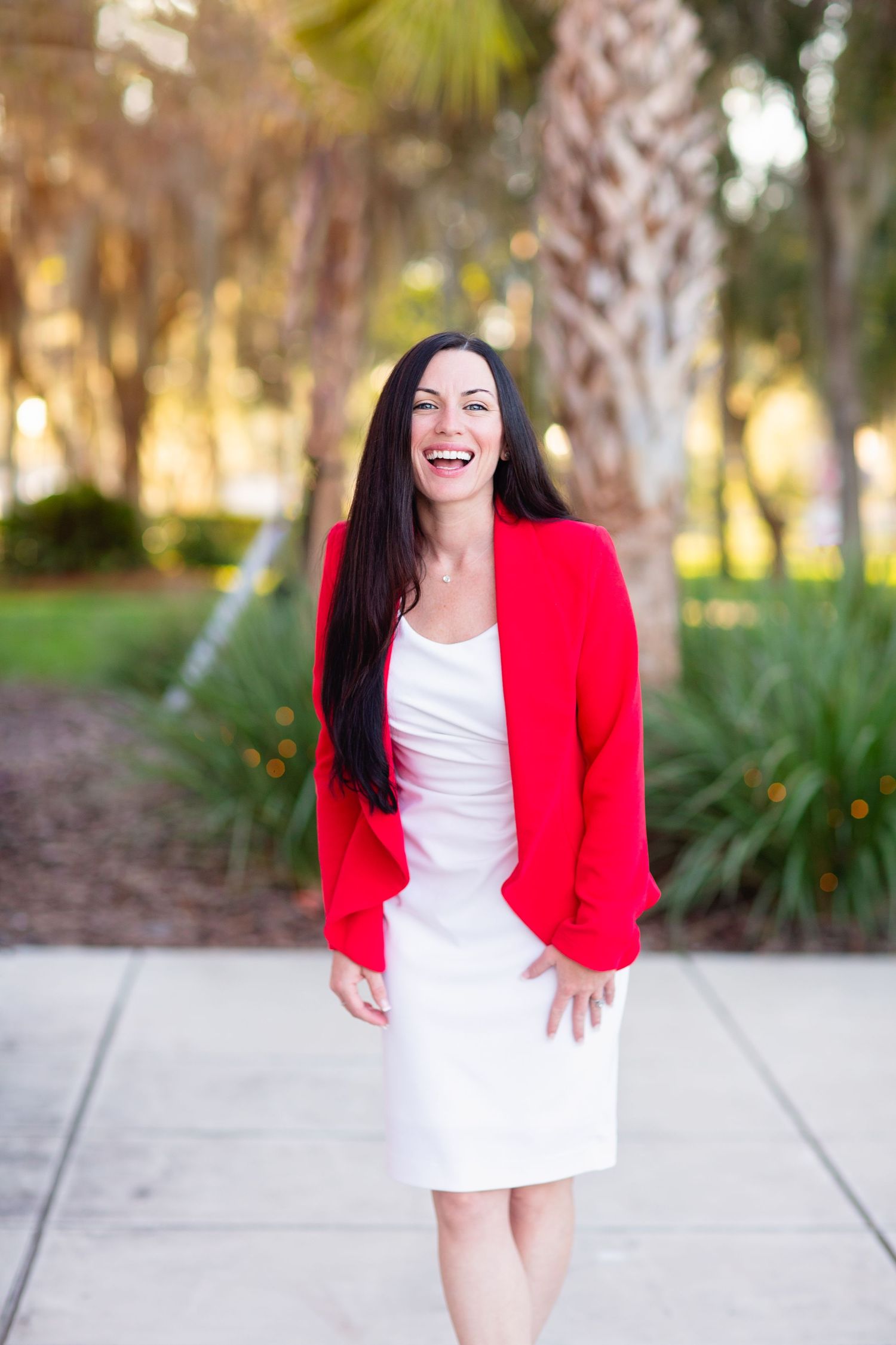 Portrait in bright red blazer and white dress showcasing joyful expression against palm trees.