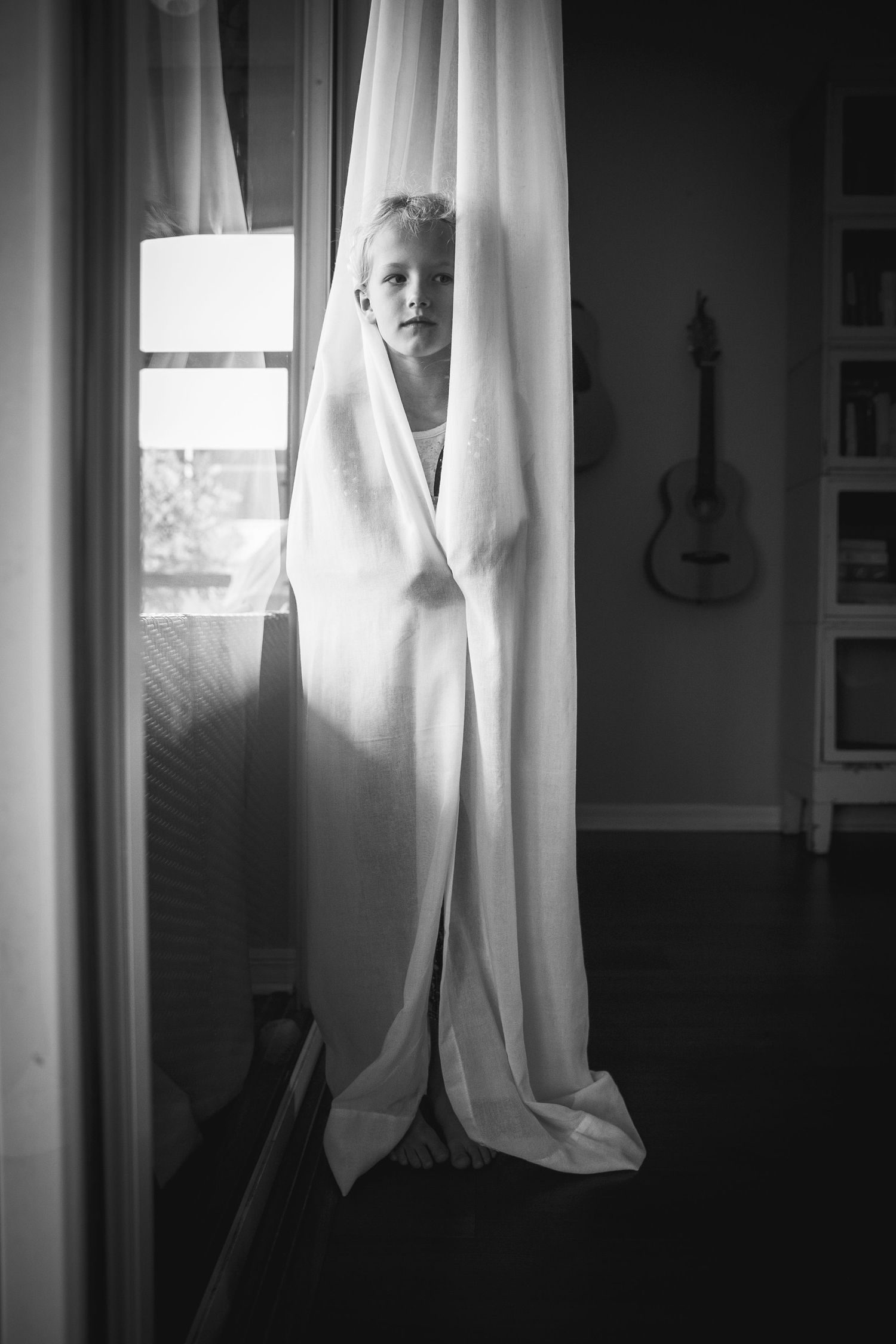 Black and white photo of child peeking through white curtains with guitar visible in background.
