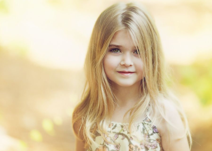 A young girl with long blonde hair poses outdoors in a floral dress with soft lighting.