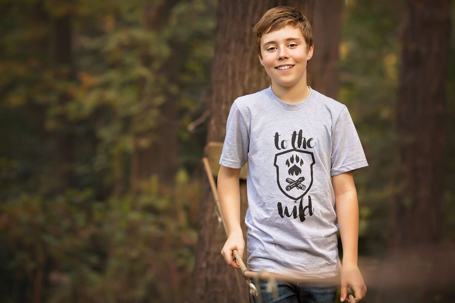 A youth wearing a gray t-shirt with a bear logo stands in front of trees in a forest setting.