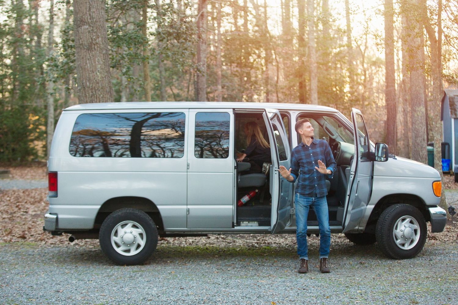 A silver van parked on a gravel path in a wooded area during golden hour sunset.