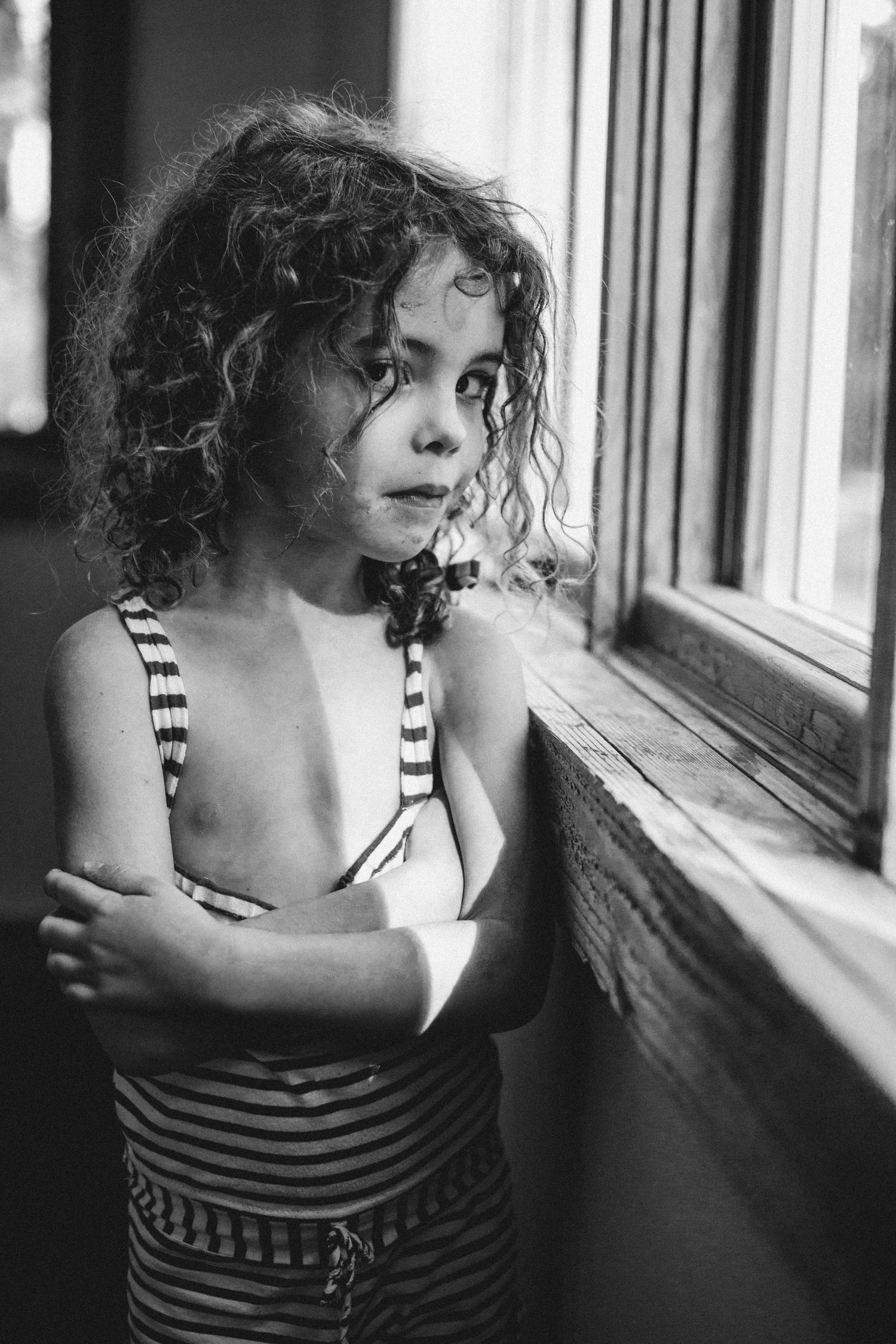 A black and white portrait of a child in striped clothing gazing thoughtfully out a window.