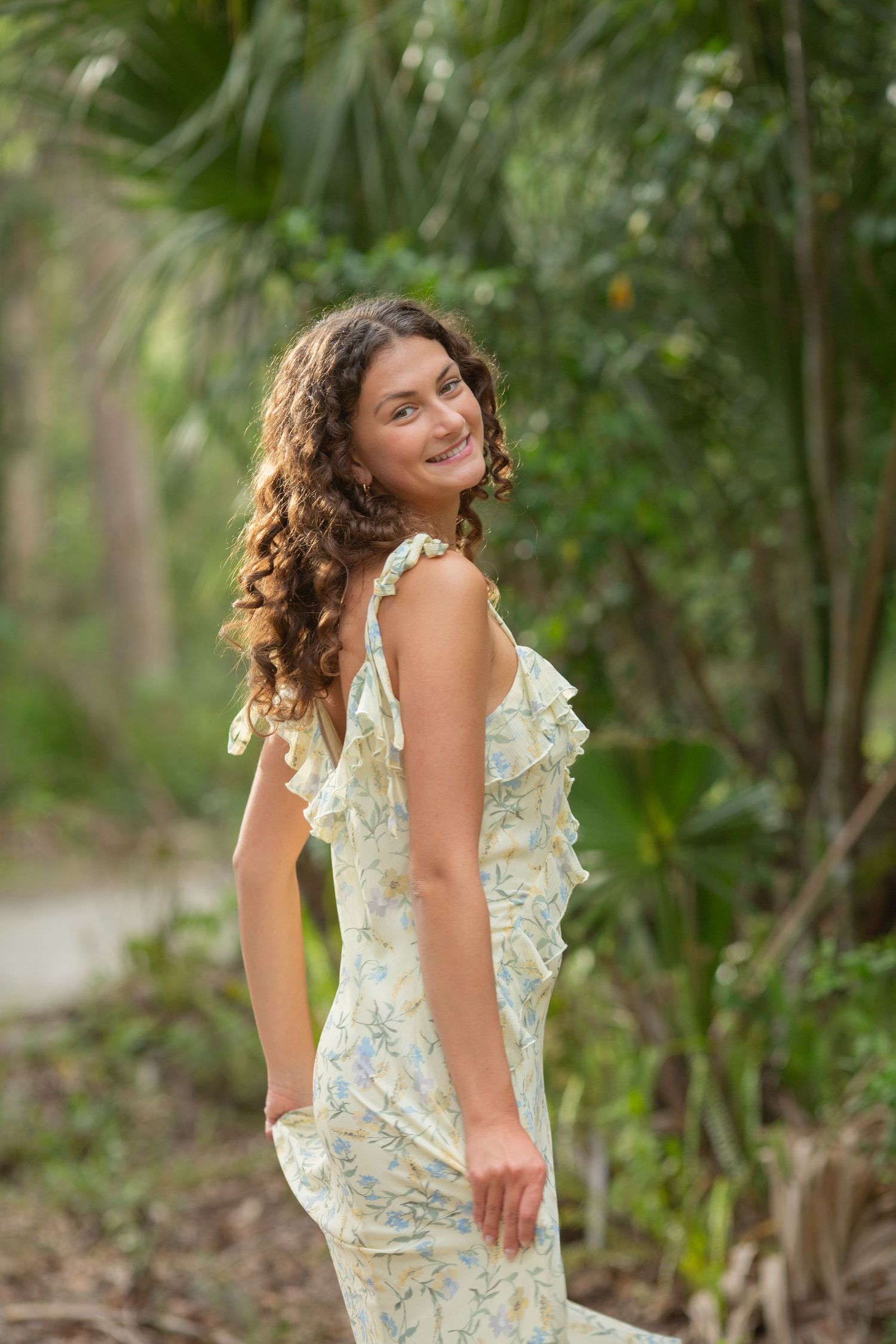 Young woman in floral sundress smiling over shoulder in lush tropical garden setting.