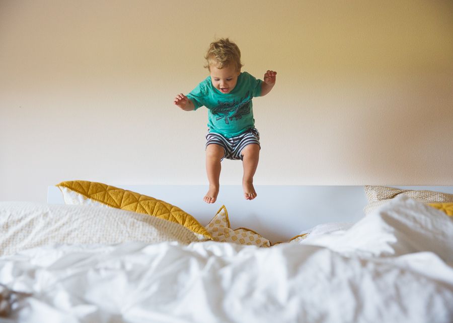 Child in teal shirt jumping playfully on white bedding.