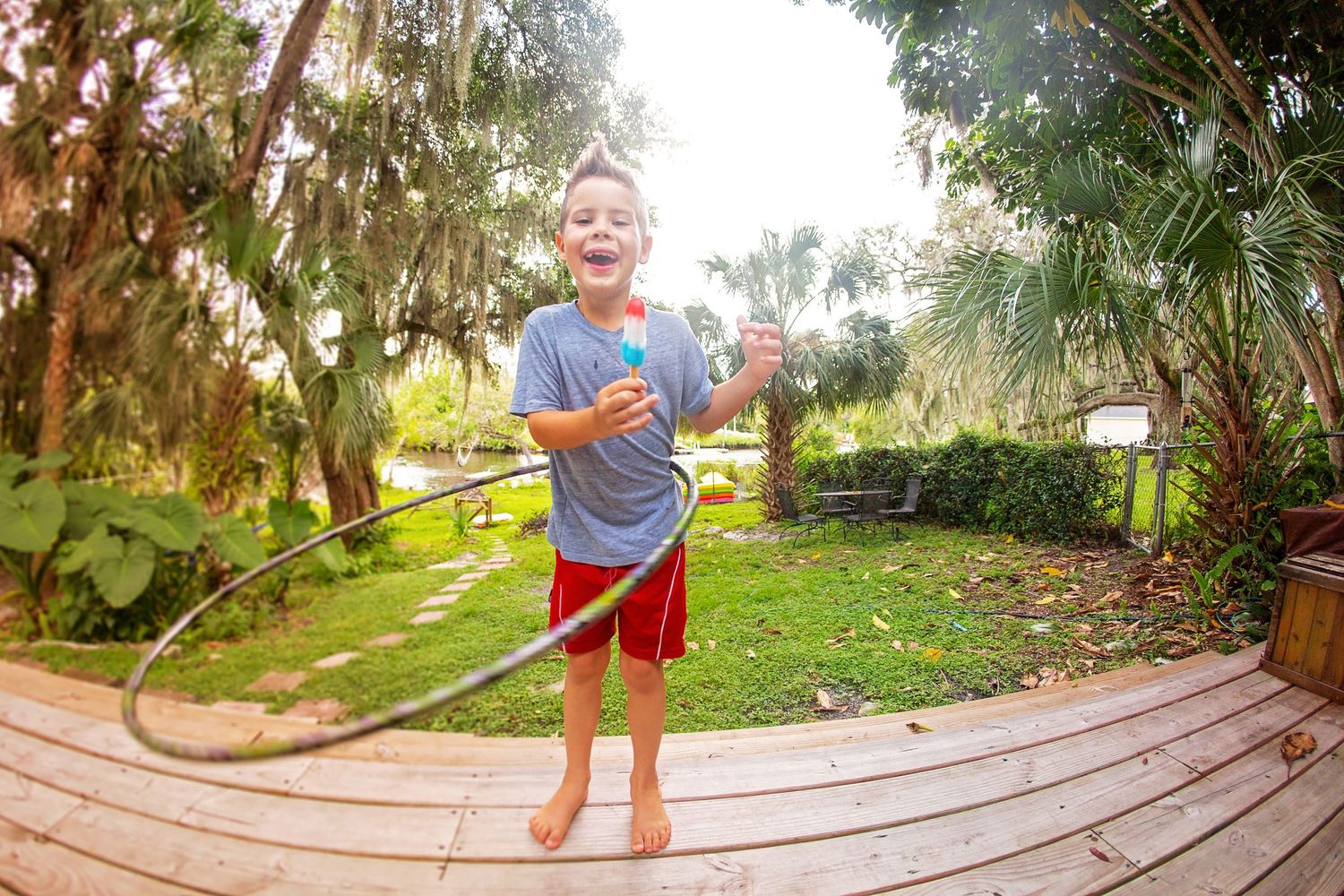 Child playing with hula hoop on wooden deck in sunny backyard.
