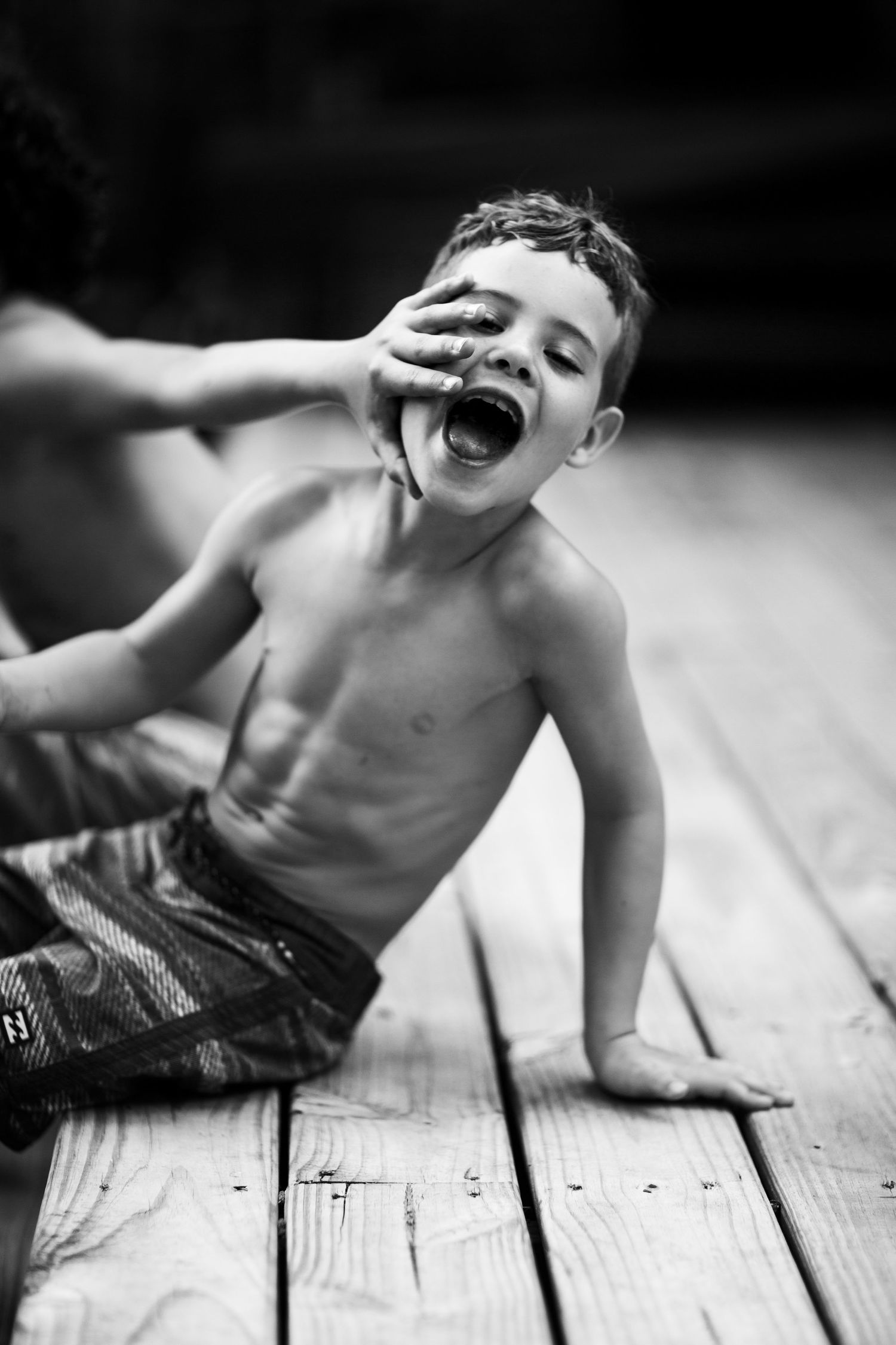 Black and white candid shot of shirtless young person laughing on wooden deck.