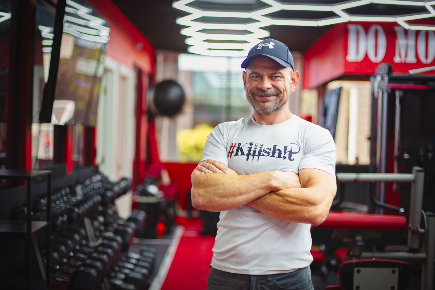 Man in white t-shirt smiling with crossed arms in red-themed fitness gym.