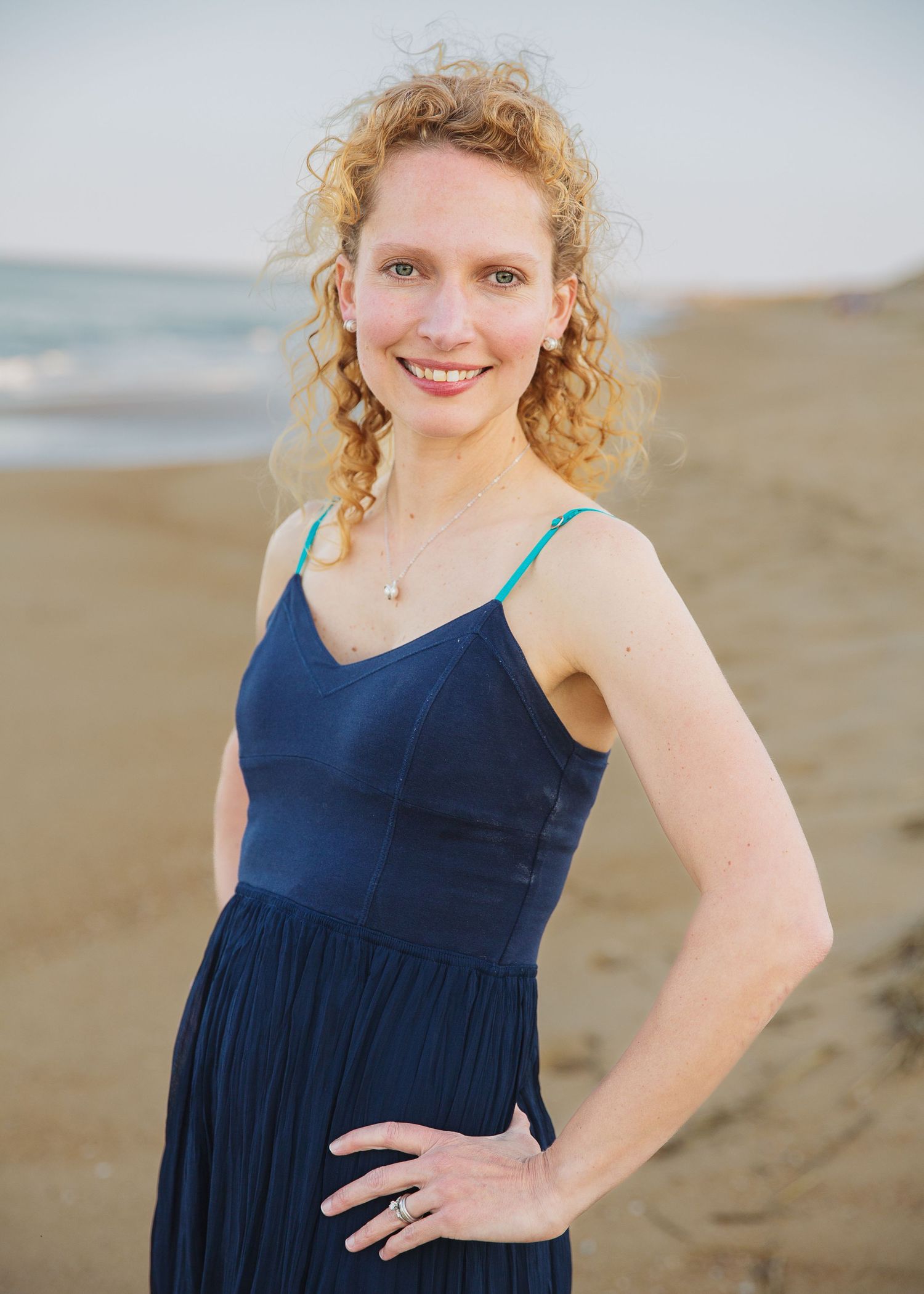 A person in a navy dress poses on a beach at sunset with natural curly hair.
