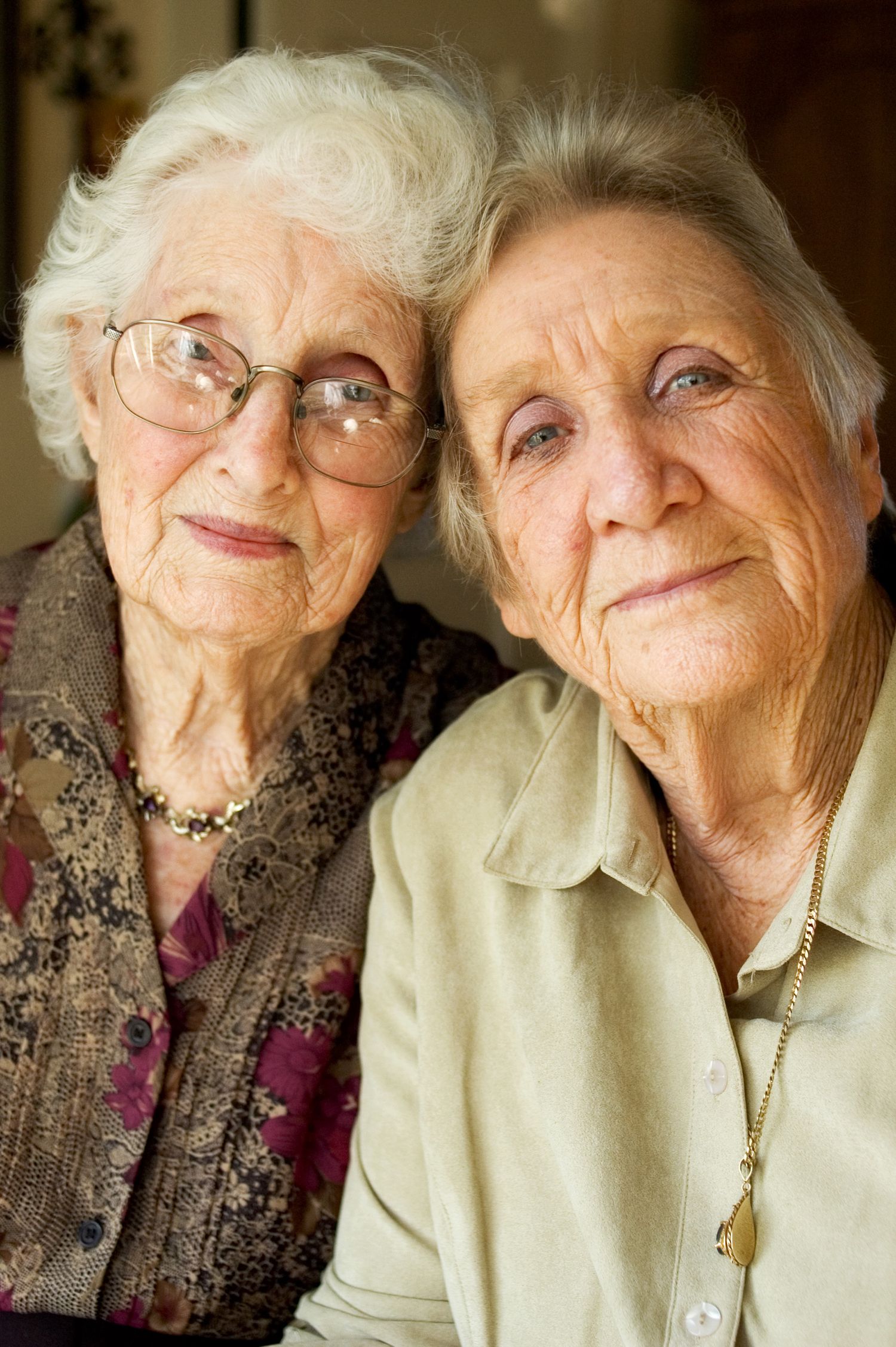 Two elderly women with glasses sit close together and smile warmly at the camera.